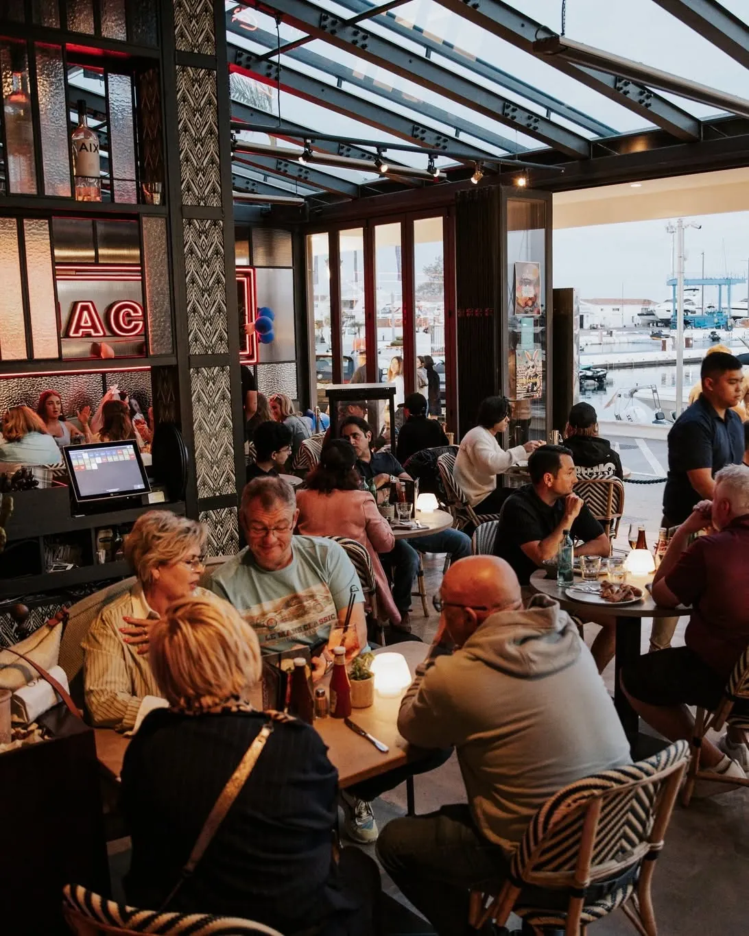 A full dining room at dusk with the marina visible through open doors