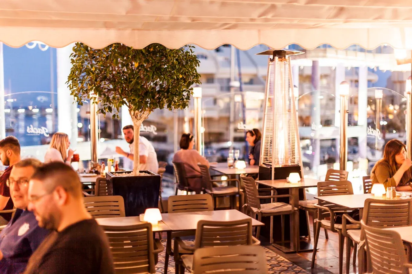 Jack's Smokehouse terrace in Benalmádena at dusk, with heaters, lamps and Puerto Marina behind