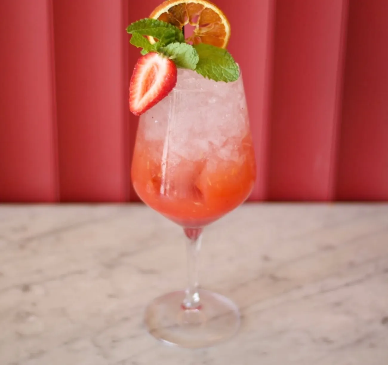 A tall strawberry spritz with mint and dried orange against a red fluted wall