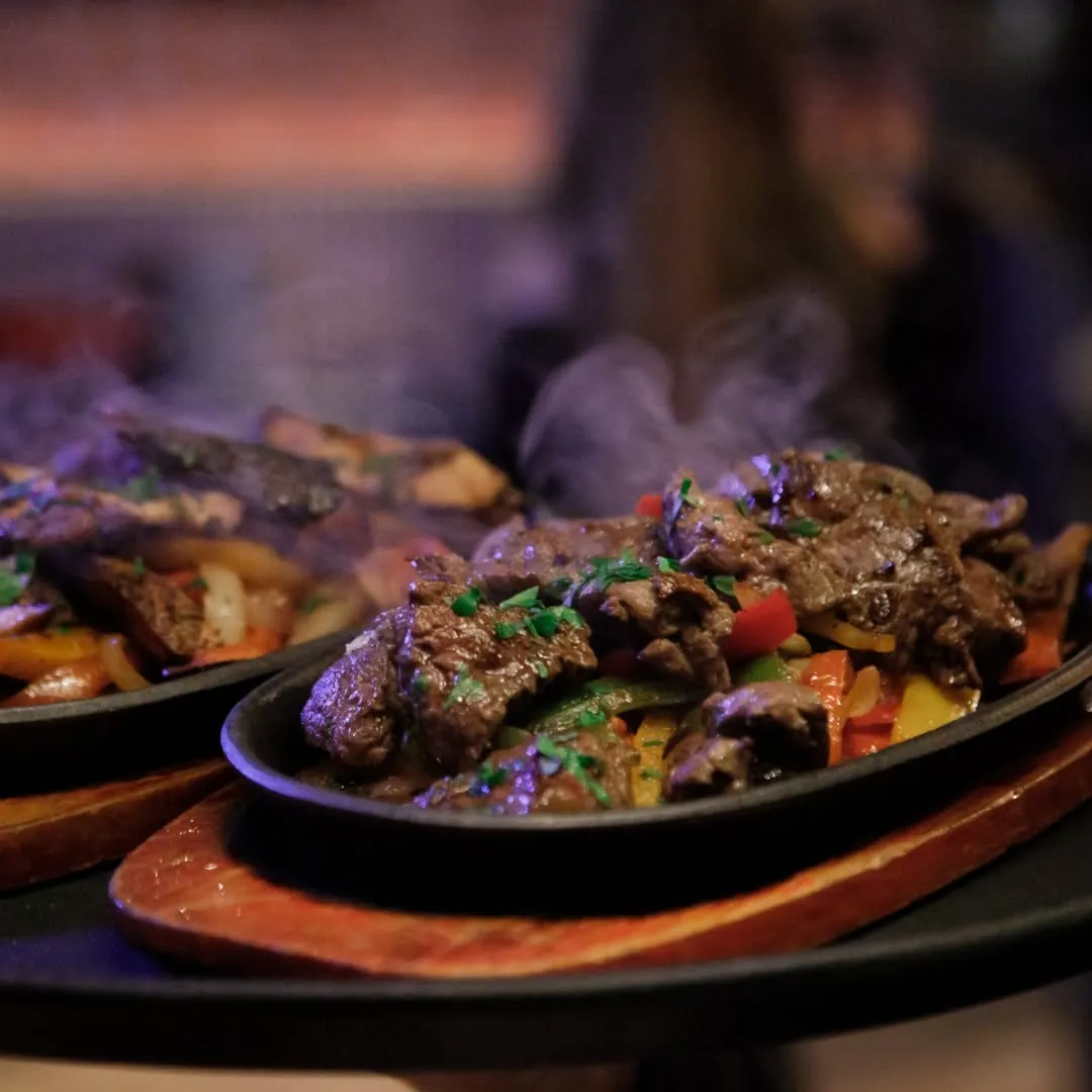Two cast-iron skillets of sizzling fajitas carried through the restaurant with smoke rising