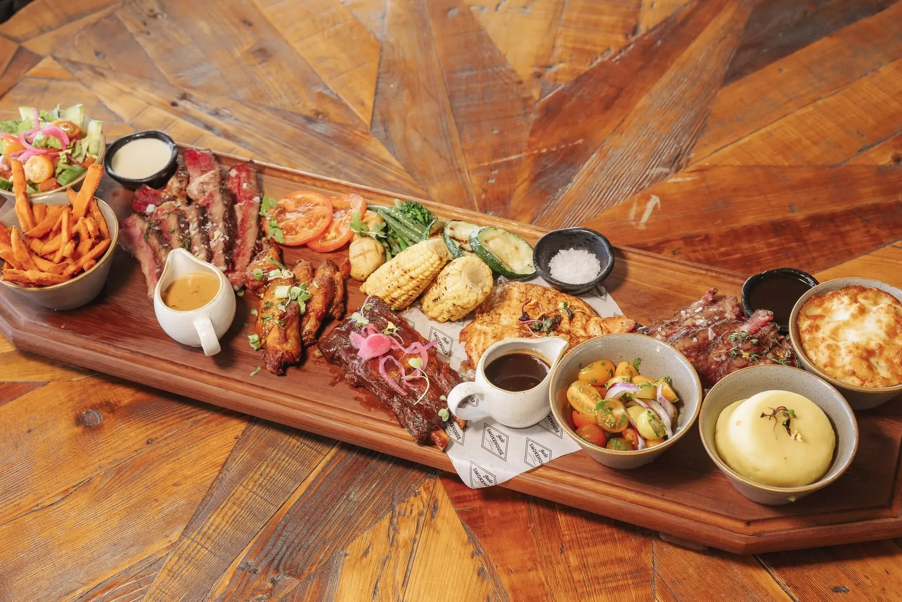 A long sharing board of ribs, steak, wings, salmon, corn and sides on a wooden table